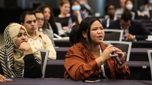 An indoor image of a participant speaking into the microphone, with more conference delegates seated in the background, listening.