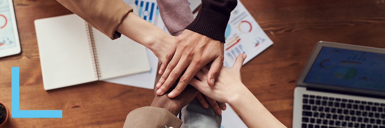 Five people reaching across an office desk, with their hands on top of one another