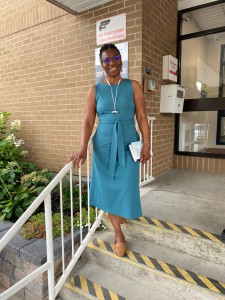 Sheila, wearing a turquoise dress, is standing by the steps at the entrance of the building where the ACT classes are.