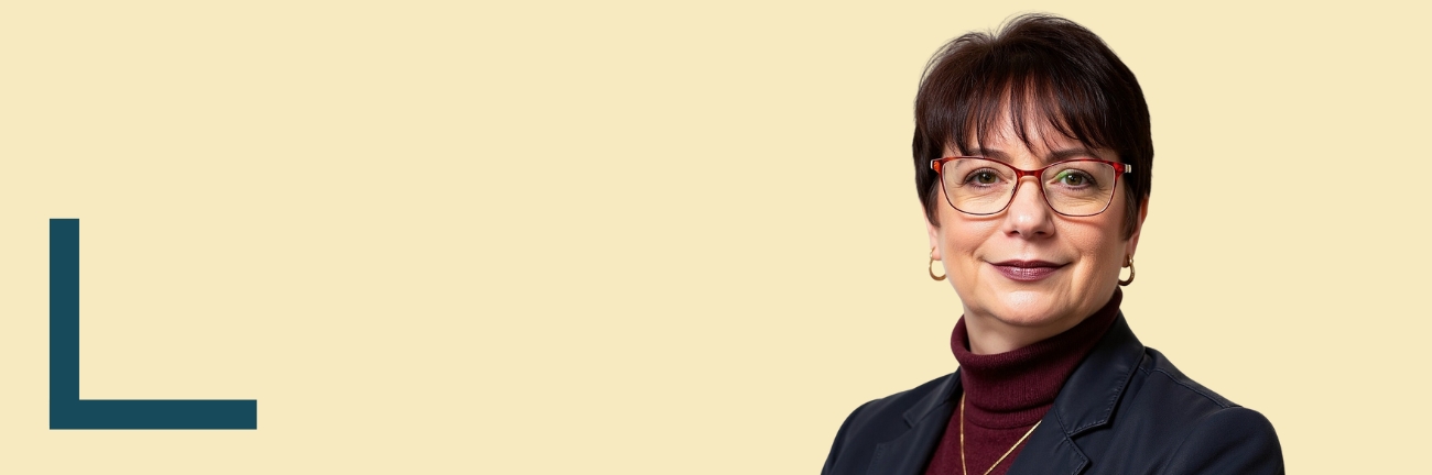 Portrait of Shannon Gill with short dark hair, wearing glasses, a burgundy turtleneck, and a navy blazer. She looks confidently at the camera, with a calm beige background.