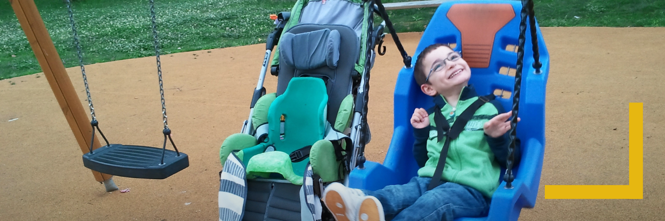 Un jeune garçon portant des lunettes, assis sur une balançoire pour fauteuil roulant, sourit joyeusement sur une aire de jeux. À proximité se trouve une balançoire standard. On aperçoit de l'herbe et un revêtement de sol sécurisé.