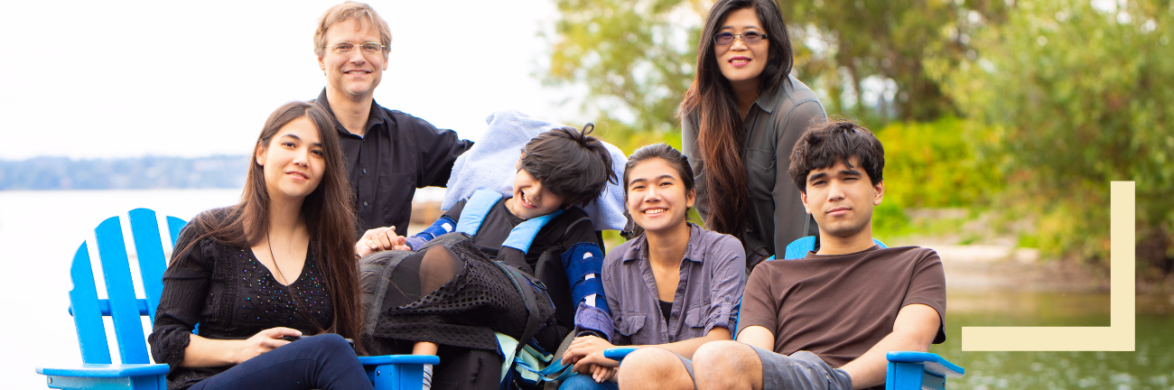 A group of six people pose happily outdoors by a lake. They are seated on vibrant blue chairs, surrounded by green trees, conveying a warm, relaxed atmosphere.