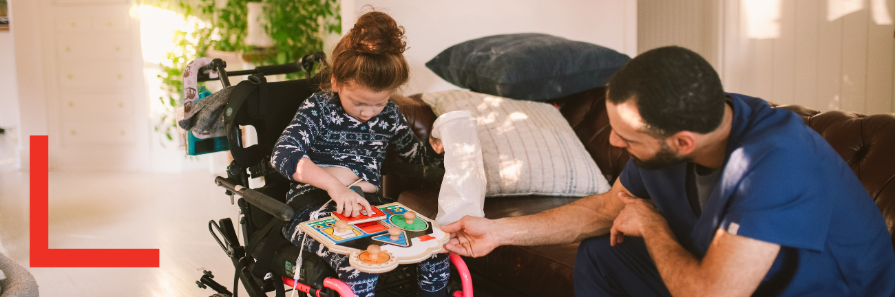 A young girl in a wheelchair plays with a puzzle, assisted by a man in blue scrubs. They are in a cozy, sunlit room with plants and a sofa.