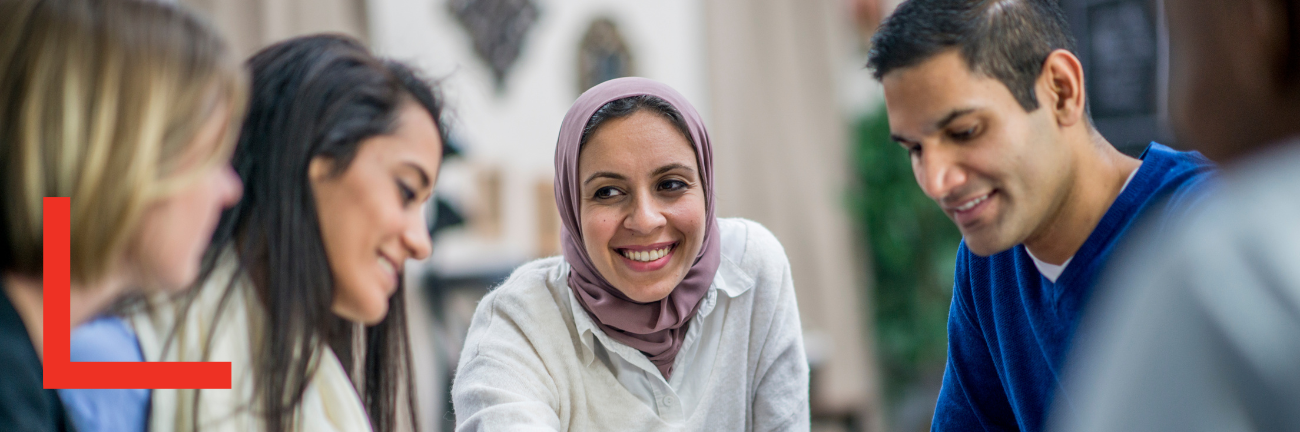 A diverse group of five people sitting and smiling, engaged in conversation. The central woman is wearing a hijab. The mood is positive and collaborative.