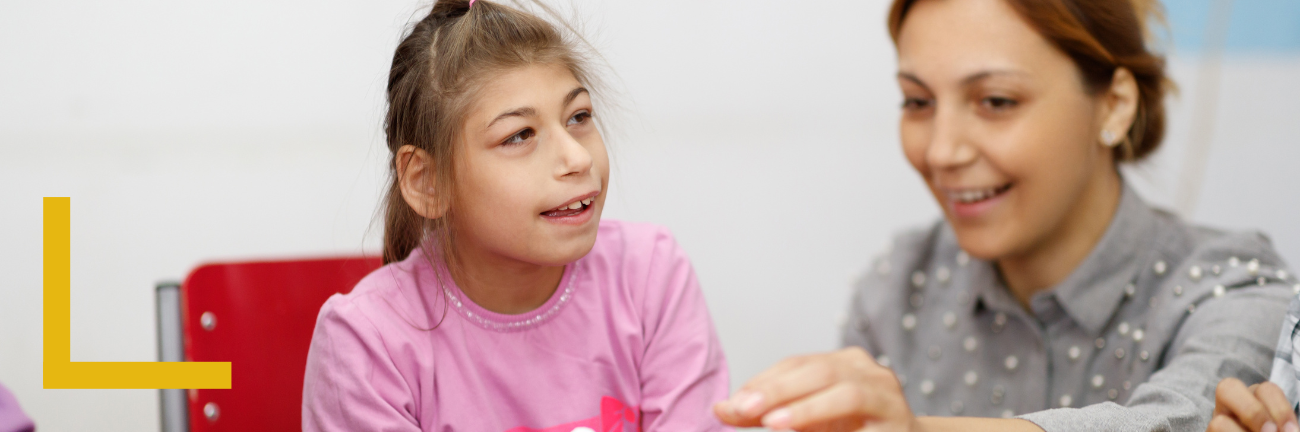 A smiling child in a pink shirt sits beside a woman with light brown hair who is gesturing with her hands. They appear engaged and happy.